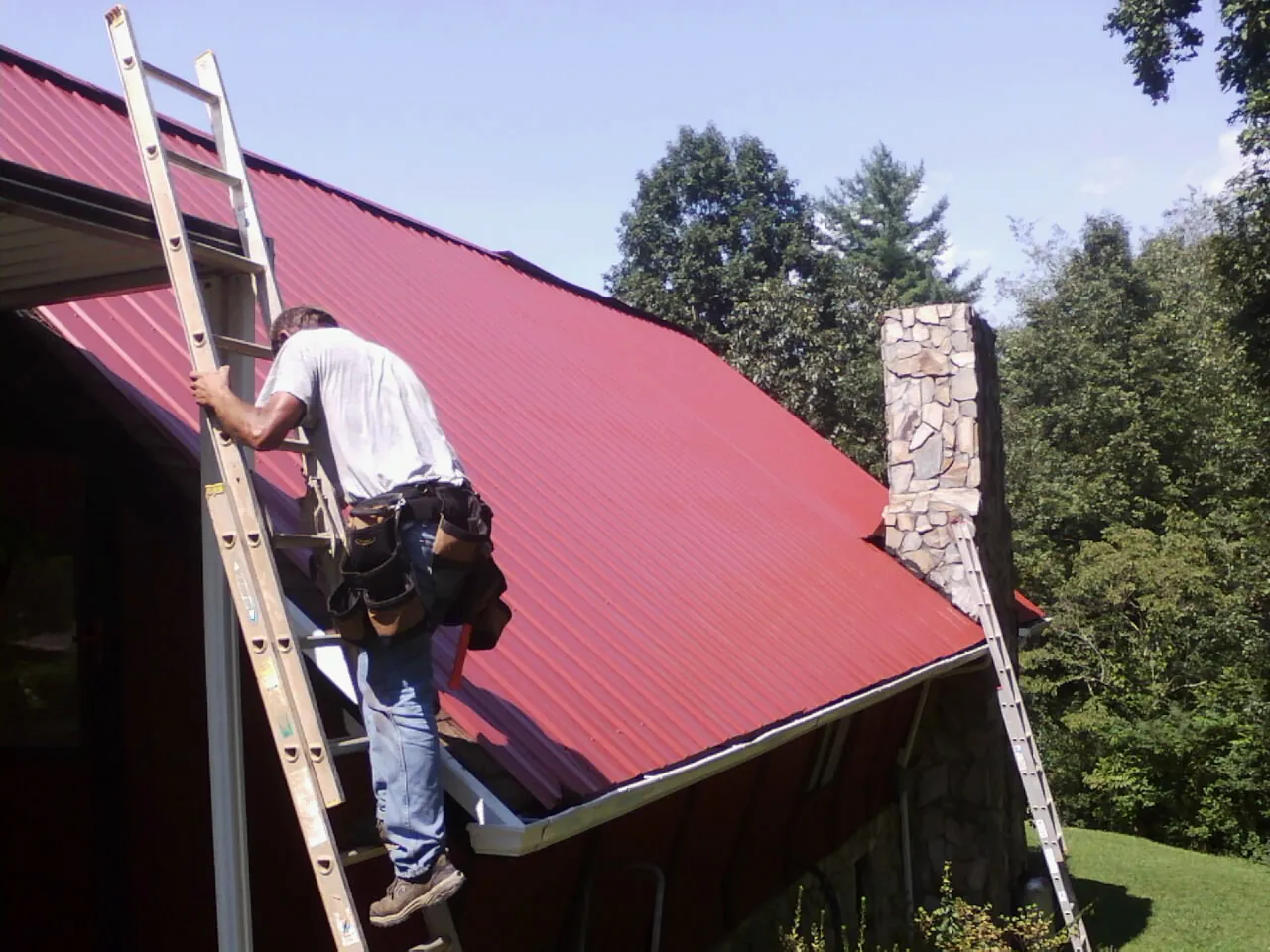 Roofer on a ladder beside a red metal roof and stone chimney — Soffit & Fascia Repair by Roofing Brothers in Green Springs